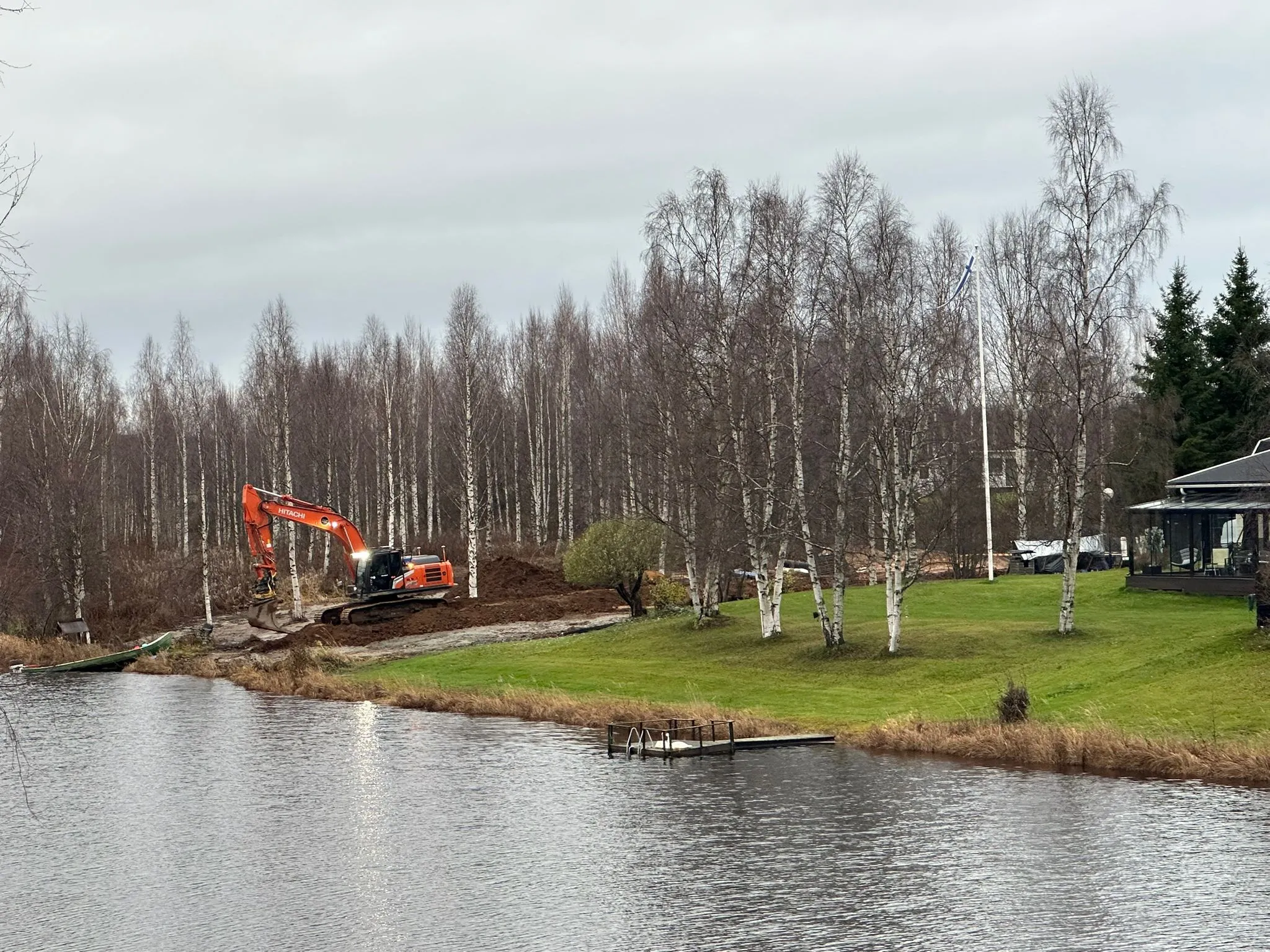 Maisema, jossa on järvi, kaivinkone, puita ja rakennus. Taivas on pilvinen ja valaistus on himmeä.
