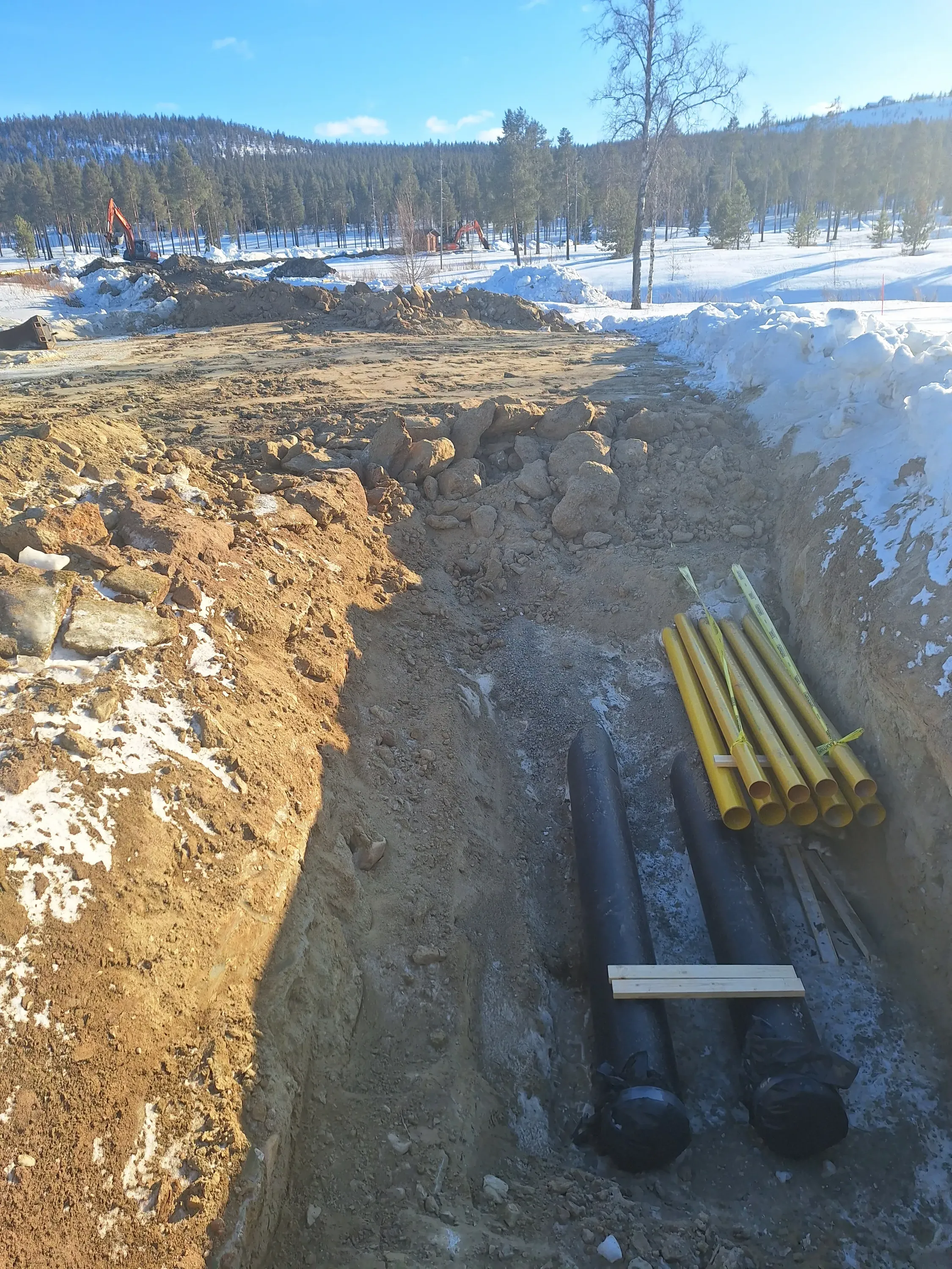 Kaivanto, jossa on mustia putkia ja nippu keltaisia putkia, maata ja lunta, aurinkoisena päivänä.