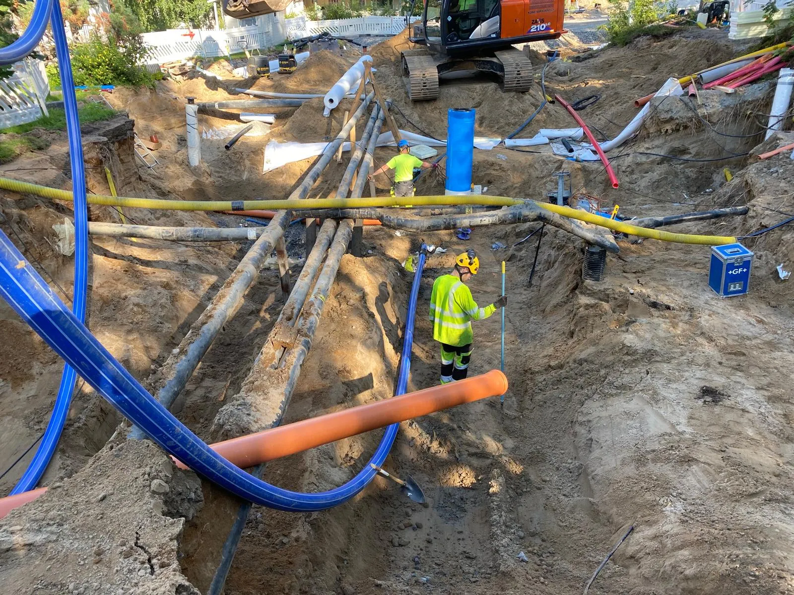 Rakennustyömaalla on kaivanto, jossa näkyy putkia, kaksi työntekijää ja kaivinkone taustalla. Työntekijöillä on suojavarusteet.