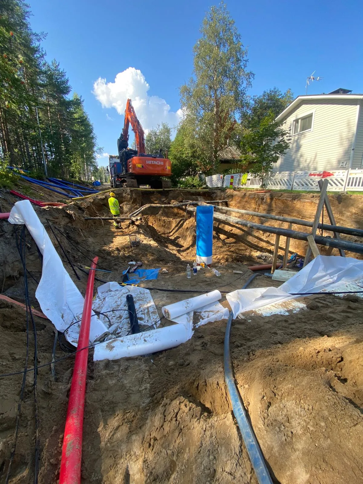 Kaivanto, jossa on putkia, kaivinkone ja työntekijä aurinkoisena päivänä.