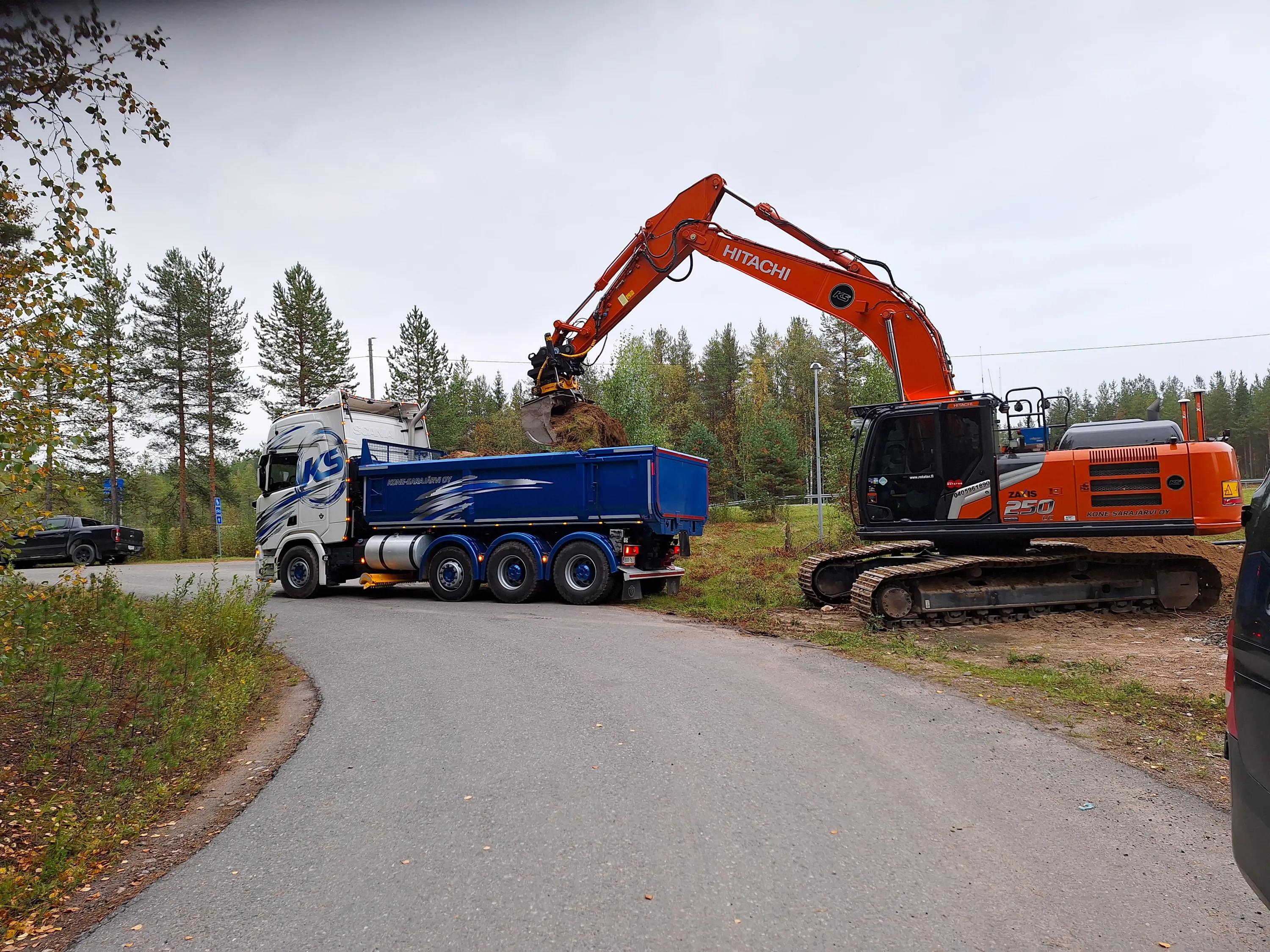 Oranssi Hitachi-kaivinkone ja sinivalkoinen kuorma-auto ovat pysäköityinä asfalttitielle. Kaivinkone on täyttämässä kuorma-auton lavan maalla. Taustalla näkyy metsää ja harmaa taivas.