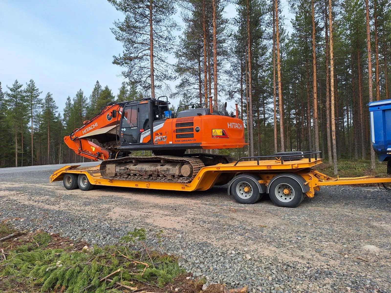 Oranssi Hitachi-kaivinkone keltaisella lavetilla metsäisellä alueella, taustalla korkeita puita ja sininen taivas.