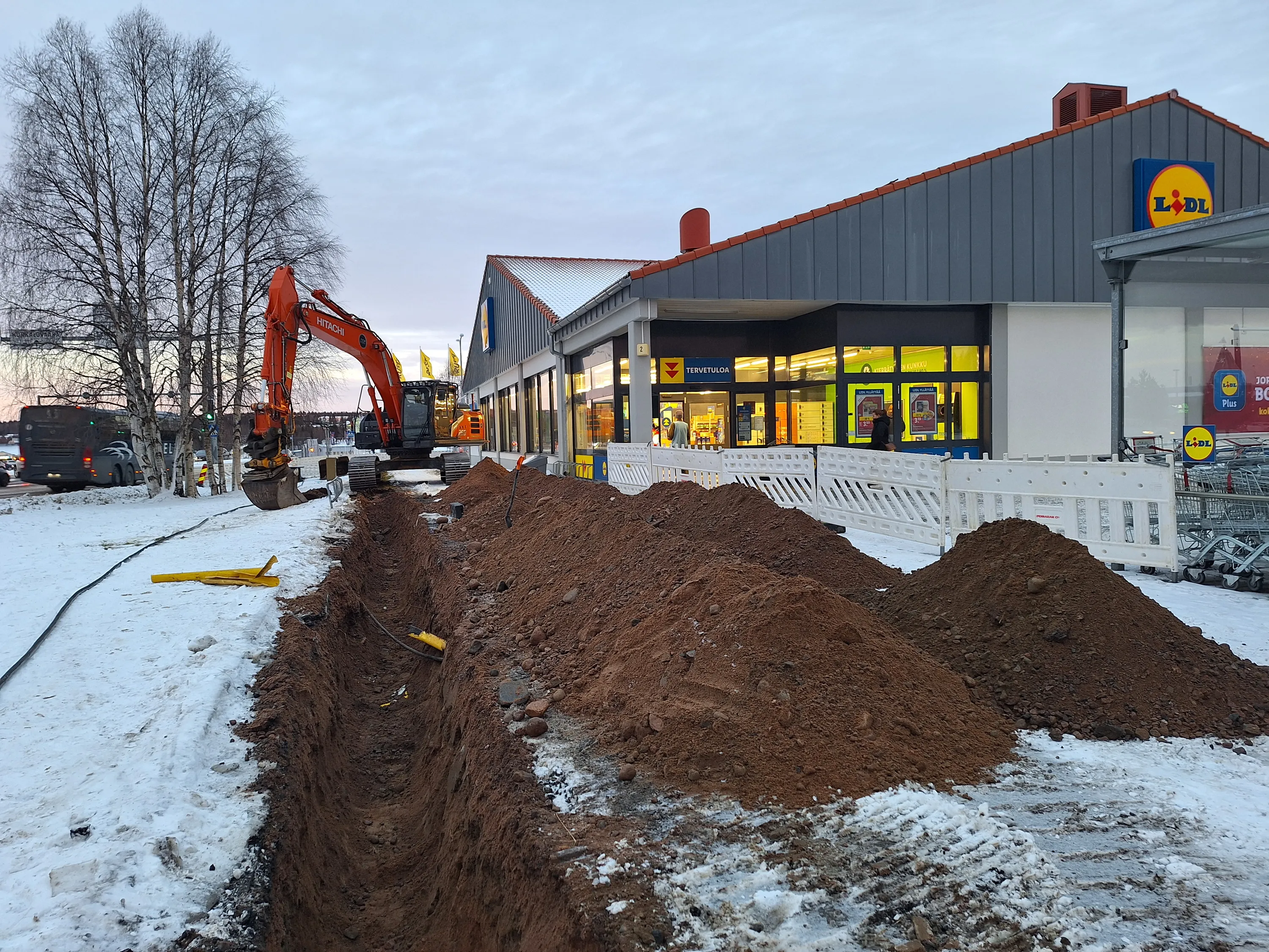 Kuvassa näkyy kaivinkone Lidl-myymälän edessä, jossa on kaivettu oja ja maata on kasattu ojan viereen. Maassa on lunta ja taivas on pilvinen.