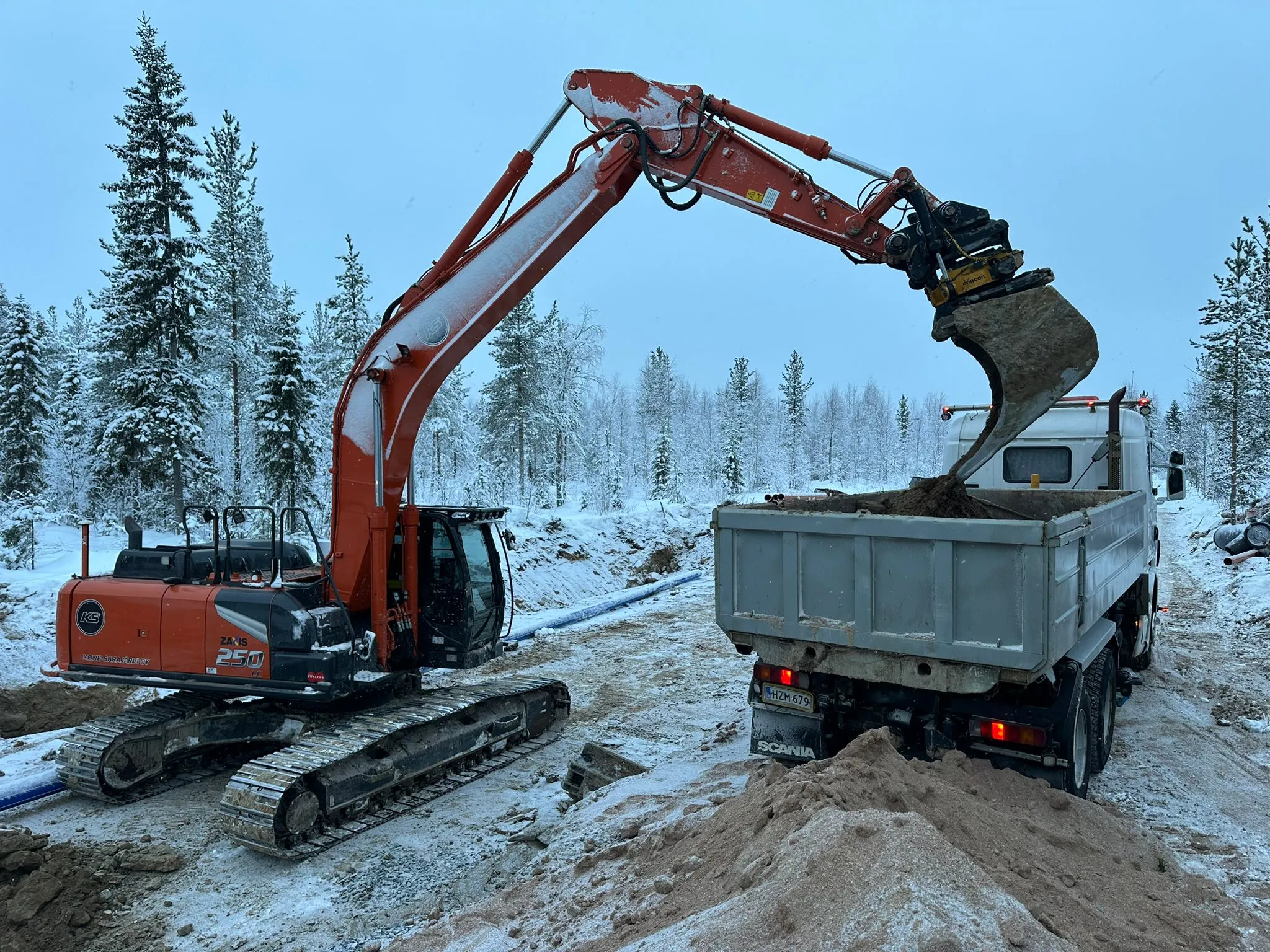 Oranssi kaivinkone täyttää harmaata kuorma-autoa lumisessa metsässä, taustalla havupuita ja harmaa taivas.