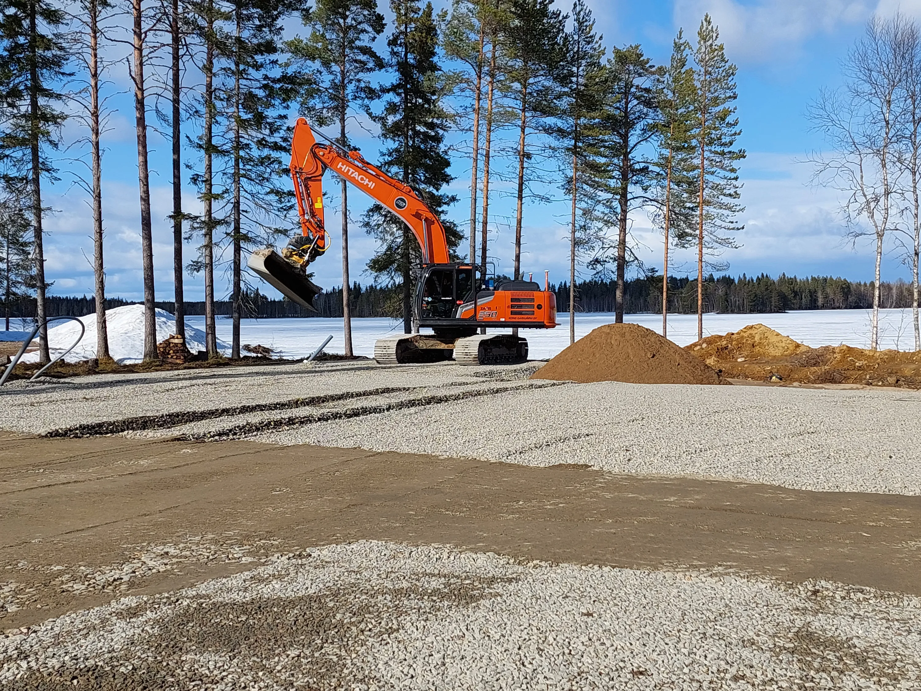Oranssi Hitachi-kaivinkone soratiellä, taustalla lumihuippuinen järvi ja havupuita aurinkoisena päivänä.