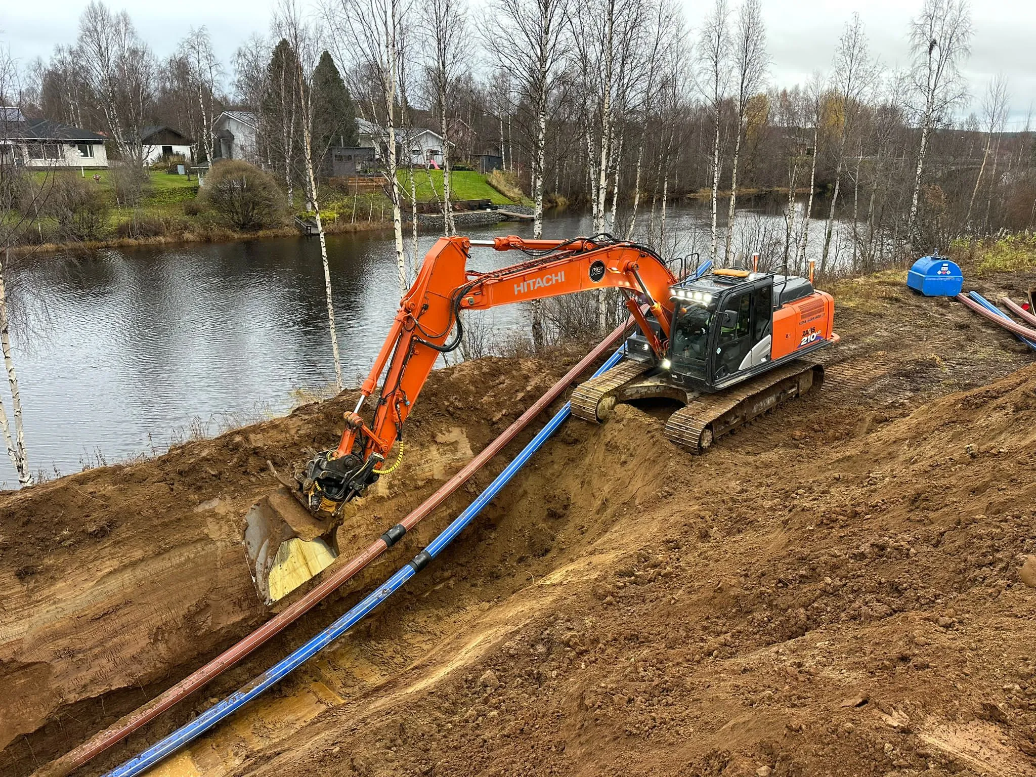 Oranssi Hitachi-kaivinkone kaivaa ojaa ruskean ja sinisen putken vierestä, taustalla järvi ja taloja pilvisessä säässä.