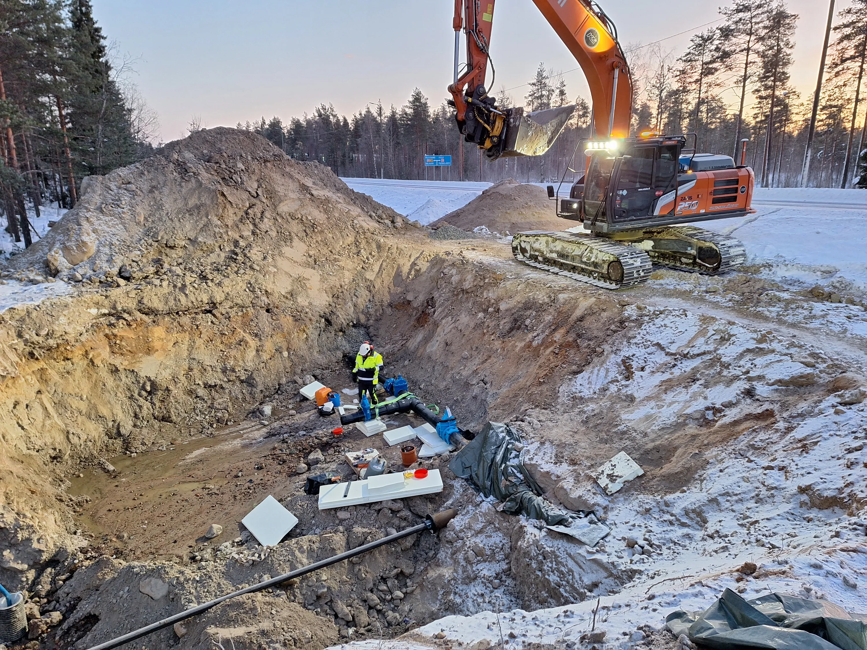 Kaivinkone kaivaa ojaa, jossa työntekijä työskentelee putkien parissa, talvinen maisema.