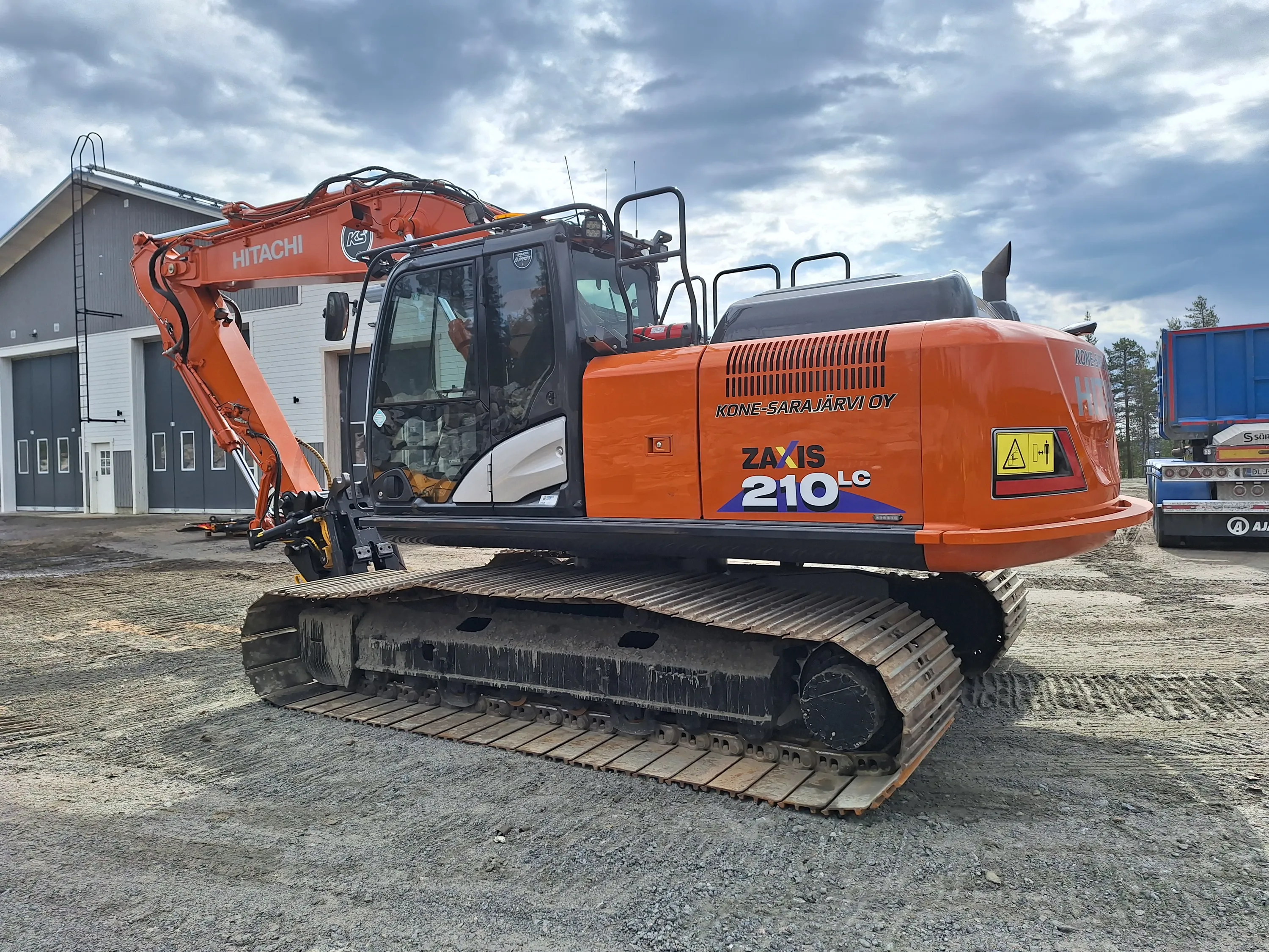 Oranssi Hitachi Zaxis 210LC -kaivinkone seisoo sorapintaisella alueella pilvisen taivaan alla. Taustalla näkyy harmaa rakennus ja sininen kuorma-auto.