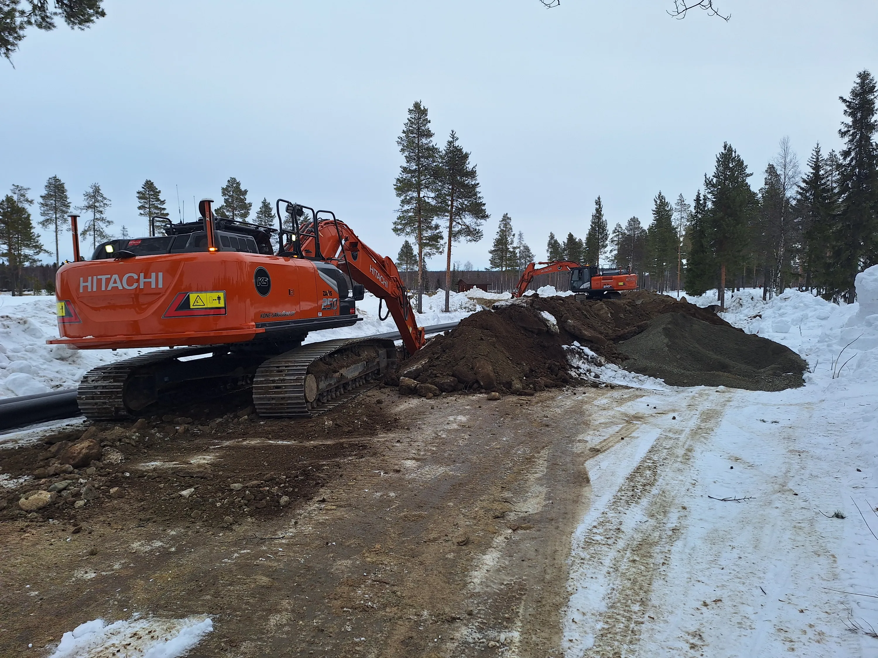 Oranssi Hitachi-kaivinkone työmaalla lumisessa metsässä, toinen kaivinkone taustalla, harmaa taivas.