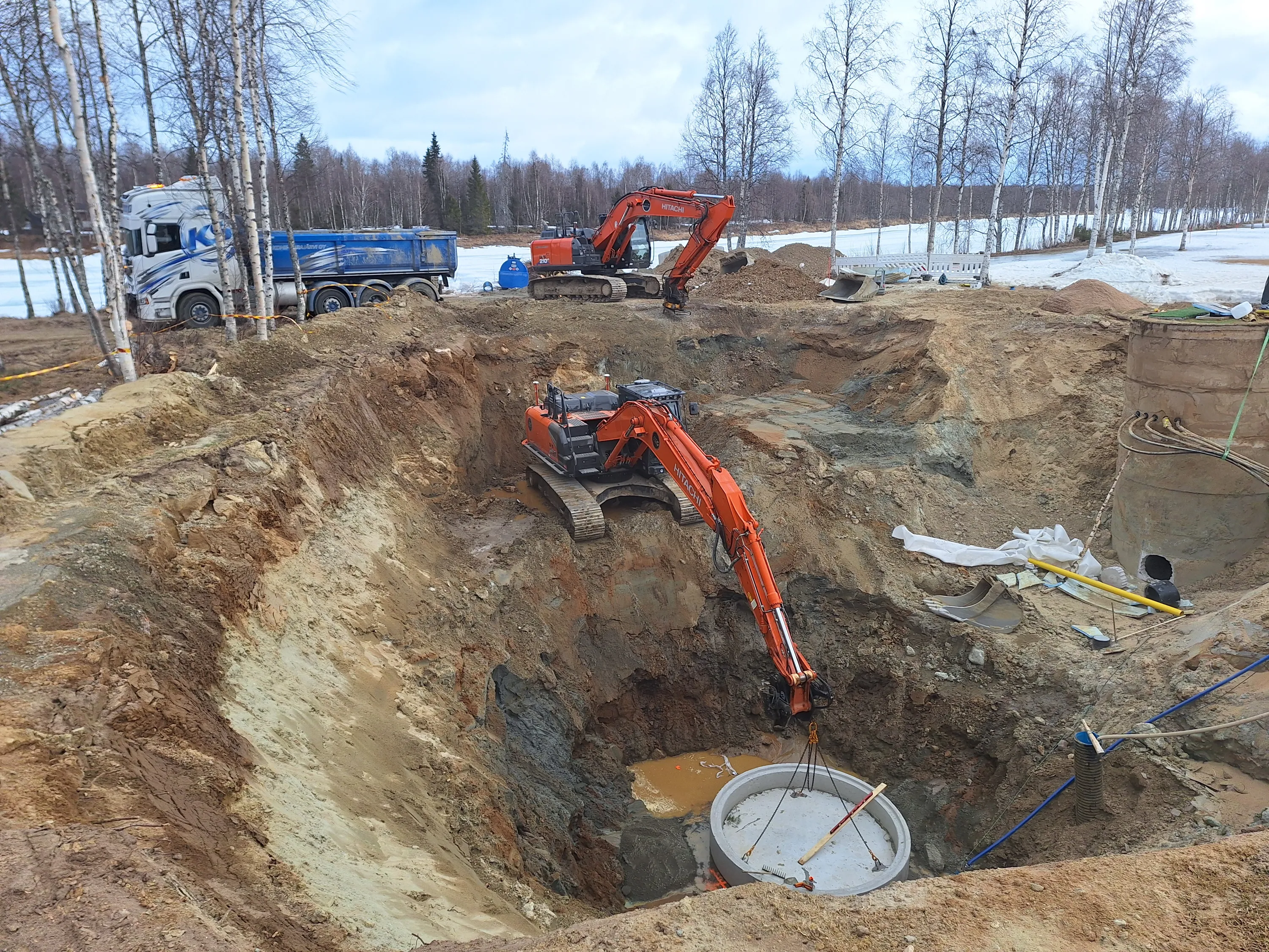 Rakennustyömaa, jossa on kaksi oranssia kaivinkonetta, kuorma-auto ja suuri kuoppa, jossa on betonielementti. Taustalla on puita ja lunta. Valoisa, mutta pilvinen sää.
