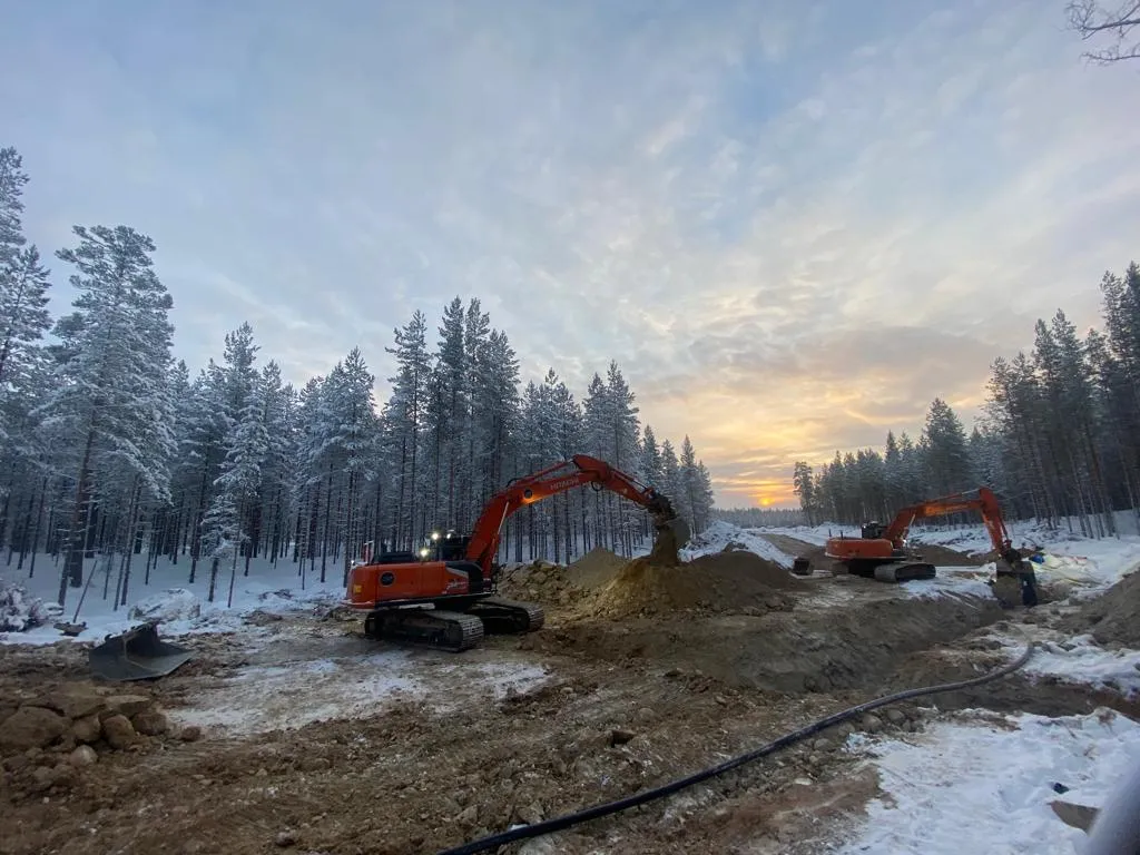 Talvinen maisema, jossa kaksi oranssia kaivinkonetta työskentelee lumisessa metsässä auringonlaskun aikaan.