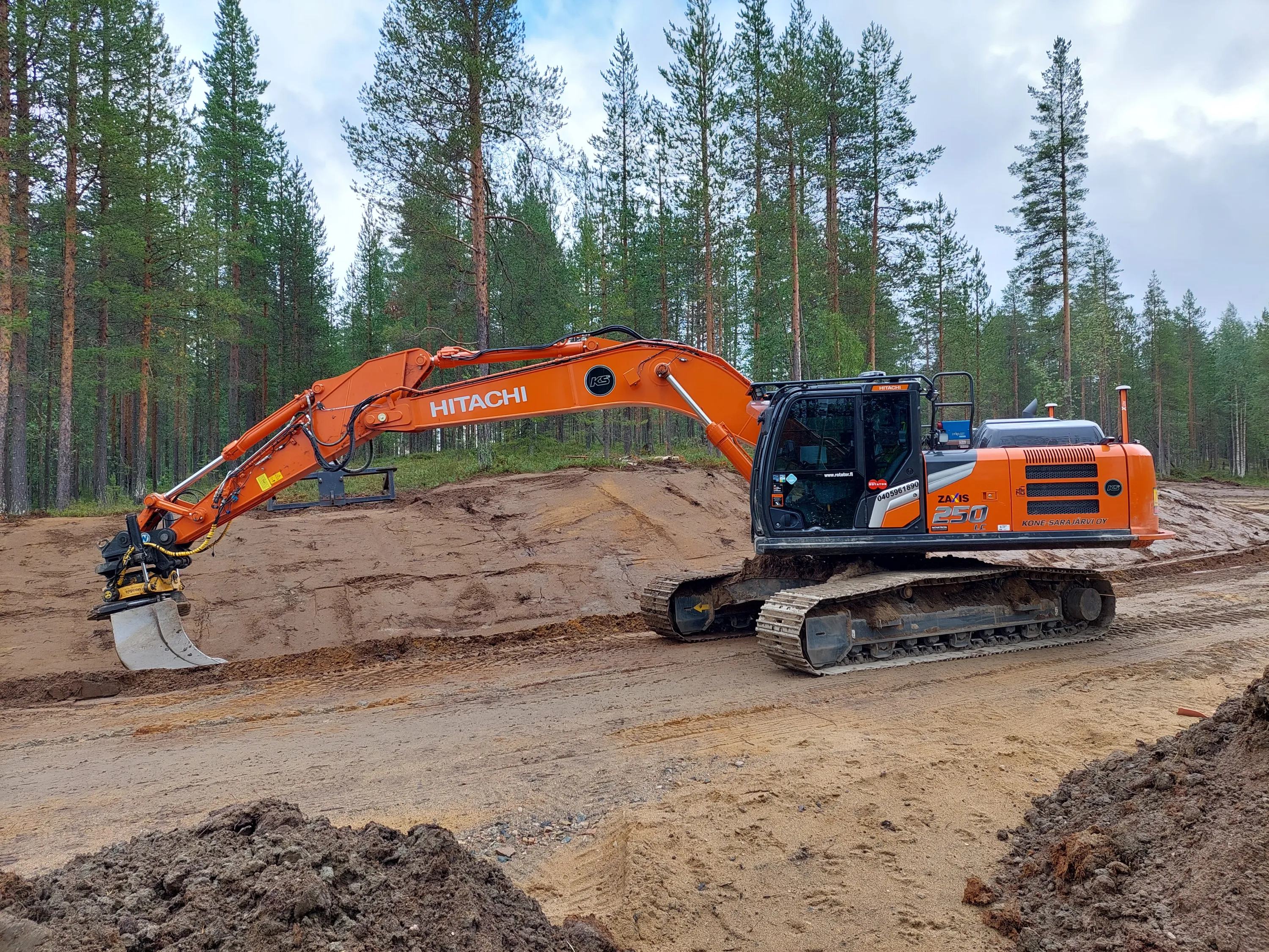 Oranssi Hitachi-kaivinkone metsäisellä työmaalla, jossa on hiekkaa ja multaa, pilvisen taivaan alla.