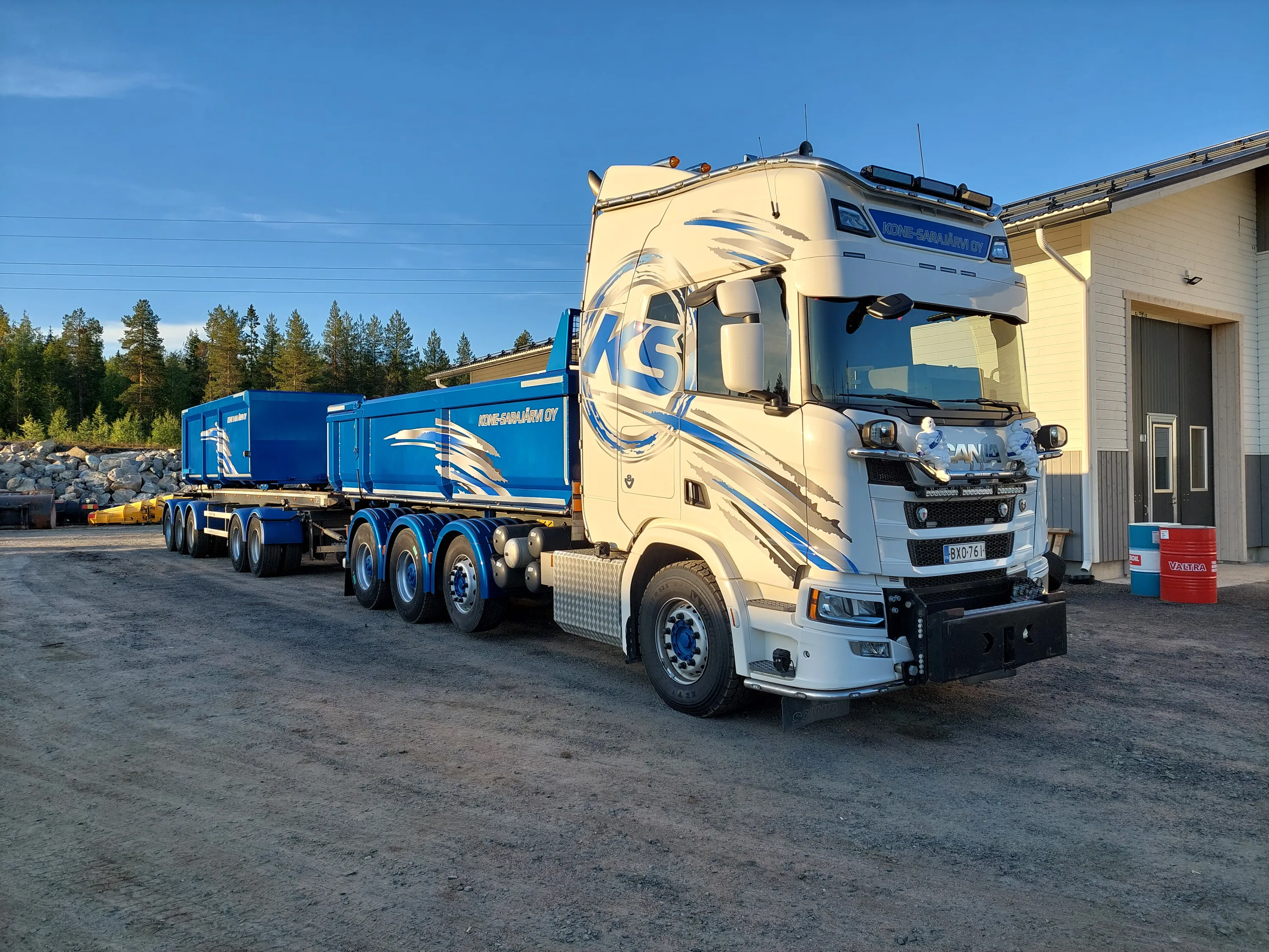 Valkoinen ja sininen kuorma-auto perävaunuineen seisoo soratiellä, taustalla metsää ja rakennus. Aurinko paistaa, valaisten kuorma-auton.