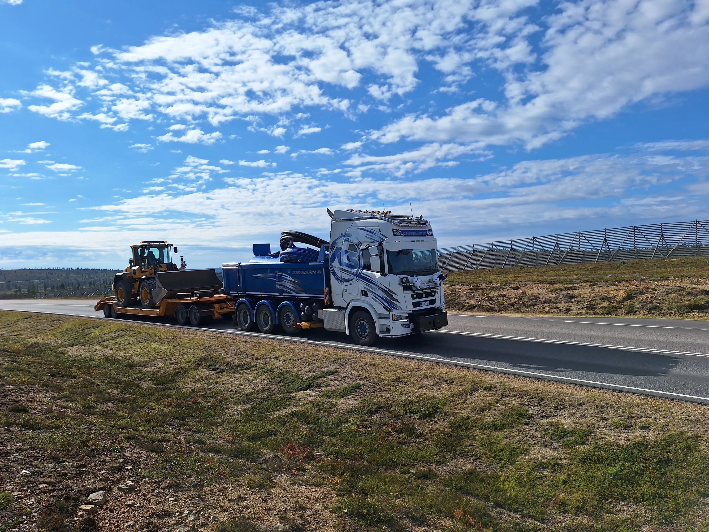 Kuorma-auto kuljettaa kaivinkonetta tiellä, taustalla sininen taivas ja pilviä.