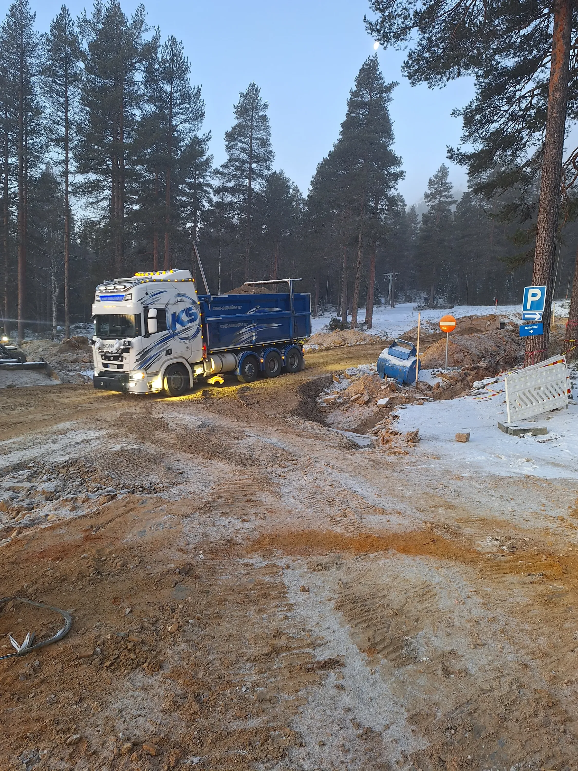 Kuorma-auto ajaa hiekkatiellä, jossa on lunta ja rakennustyömaa, taustalla metsä ja sininen taivas.