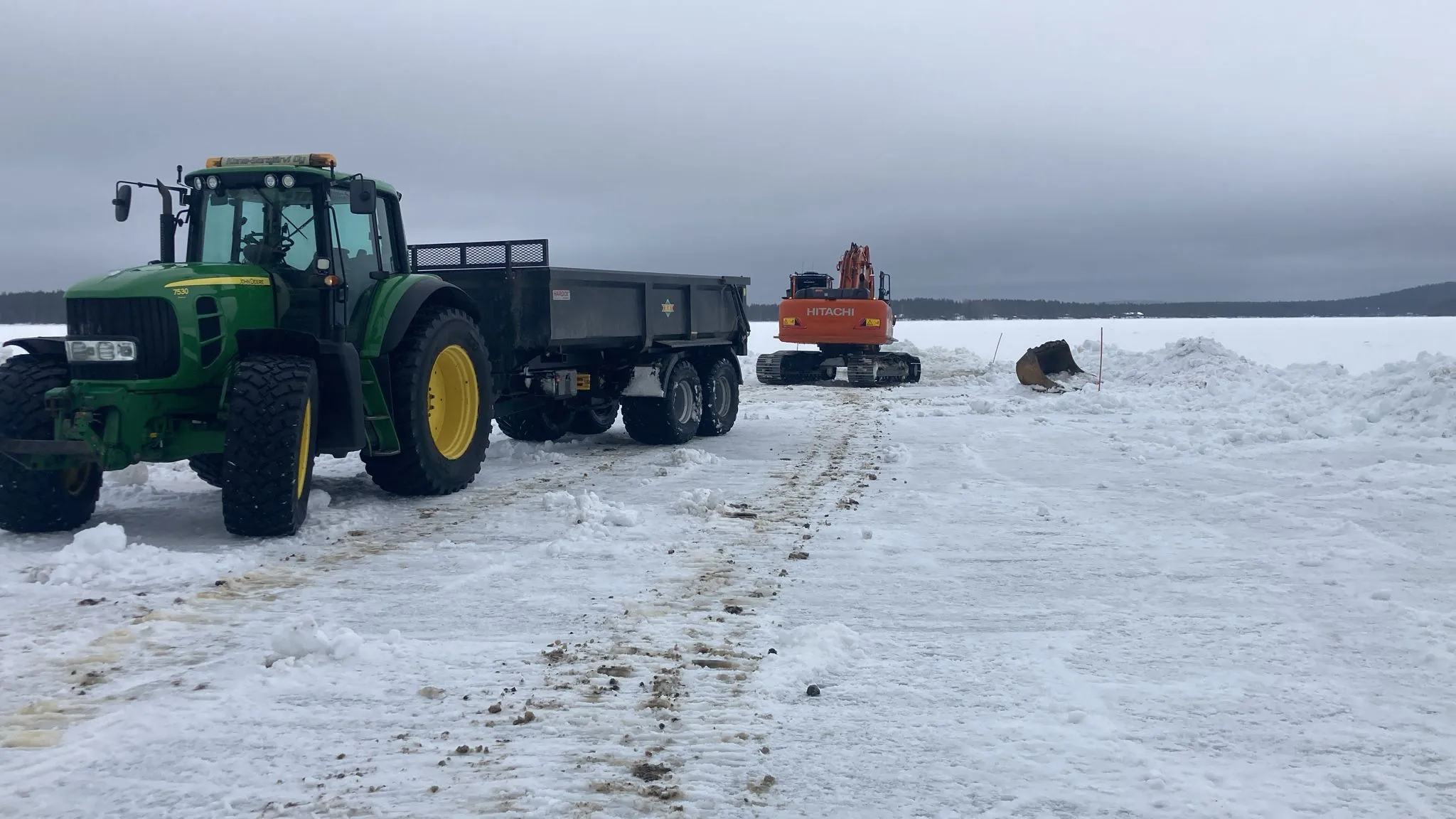 Lumisella pellolla on vihreä traktori peräkärryineen ja oranssi kaivinkone. Taustalla näkyy lumikasoja ja harmaa taivas.