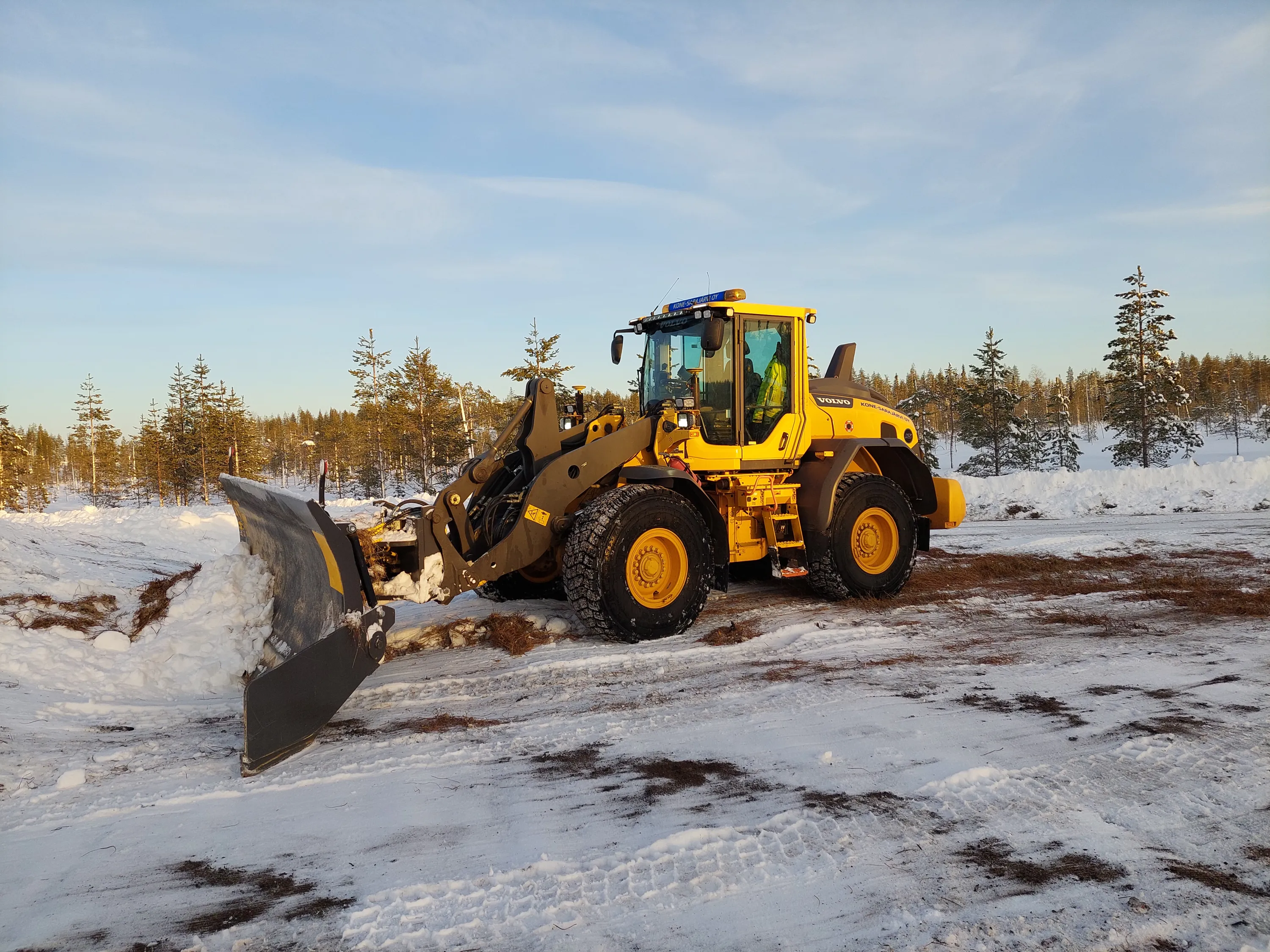 Keltainen Volvo-lumiaura auratulla lumisella tiellä, taustalla havumetsä ja sininen taivas.