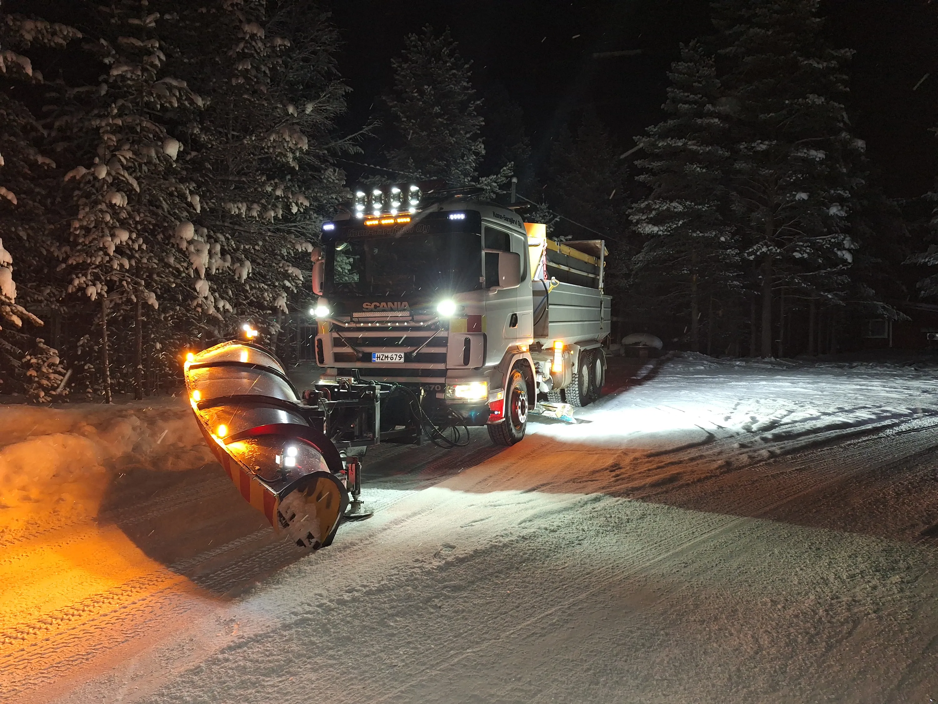 Lumiaura-auto auraa lumista tietä yöllä, valot päällä, metsän reunalla.