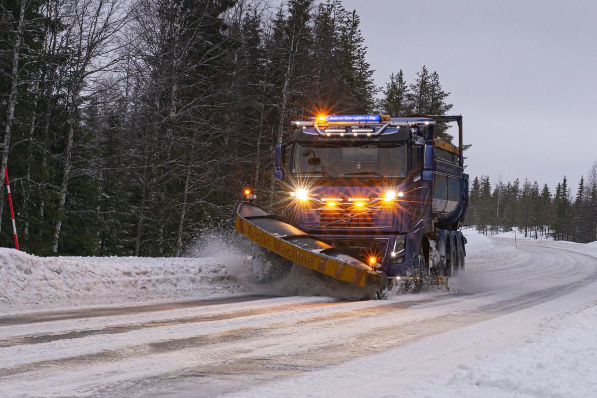 Lumiaurausauto auraa lunta lumisella tiellä metsäisessä maisemassa, valot päällä.