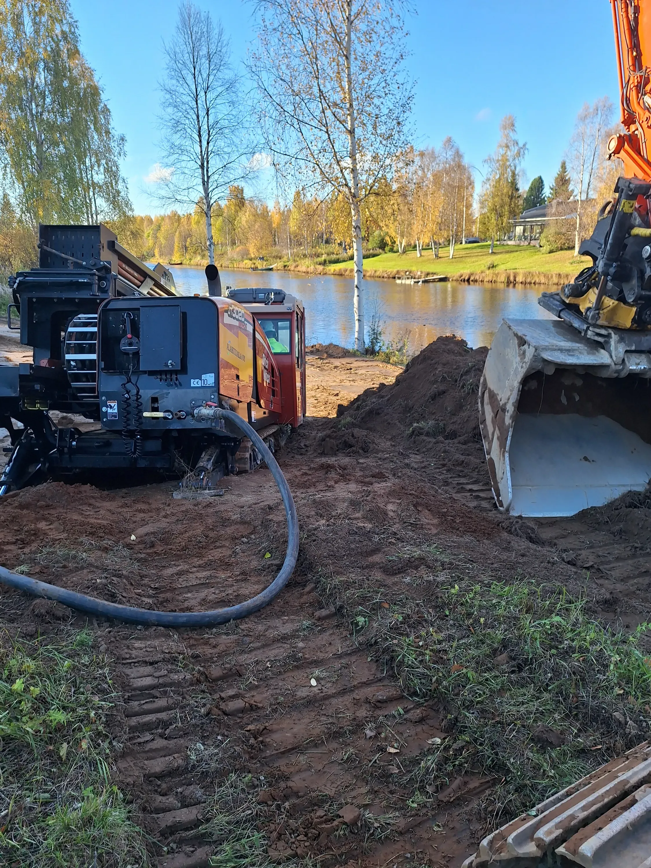Maansiirtotyömaa joen rannalla, jossa näkyy kaivinkone ja porauslaite aurinkoisena päivänä.