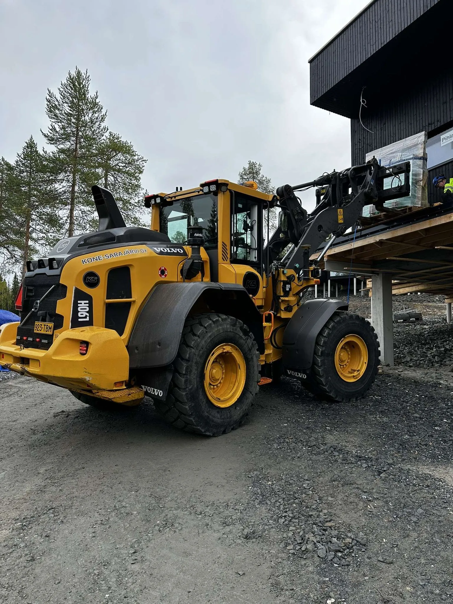 Keltainen Volvo L90H pyöräkuormaaja seisoo soratiellä rakennustyömaalla, nostaen kuorman rakennuksen päälle pilvisen sään alla.