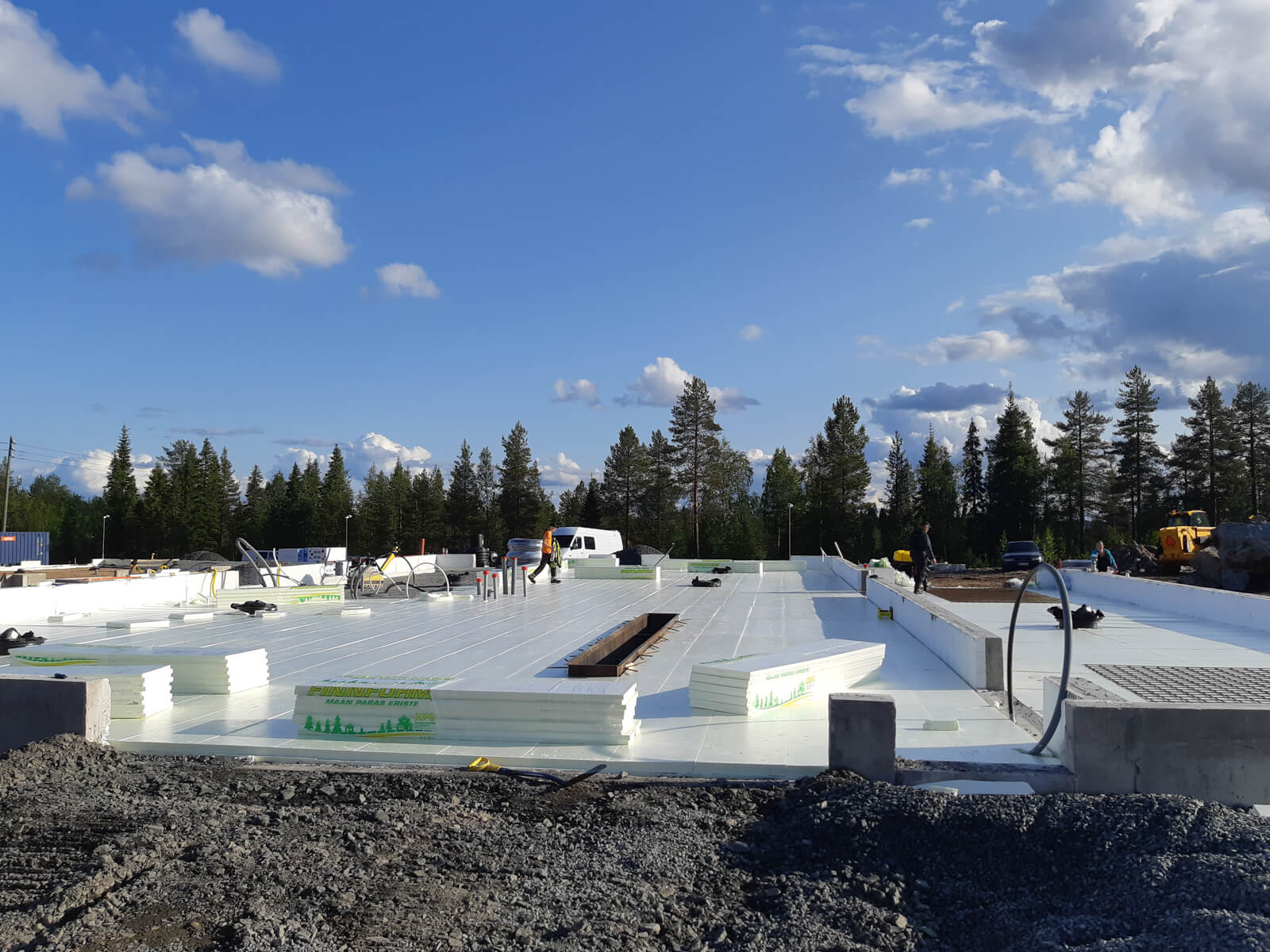 Rakennustyömaa aurinkoisena päivänä, jossa näkyy valkoista eristysmateriaalia, työntekijöitä ja taustalla metsää sinisen taivaan alla.