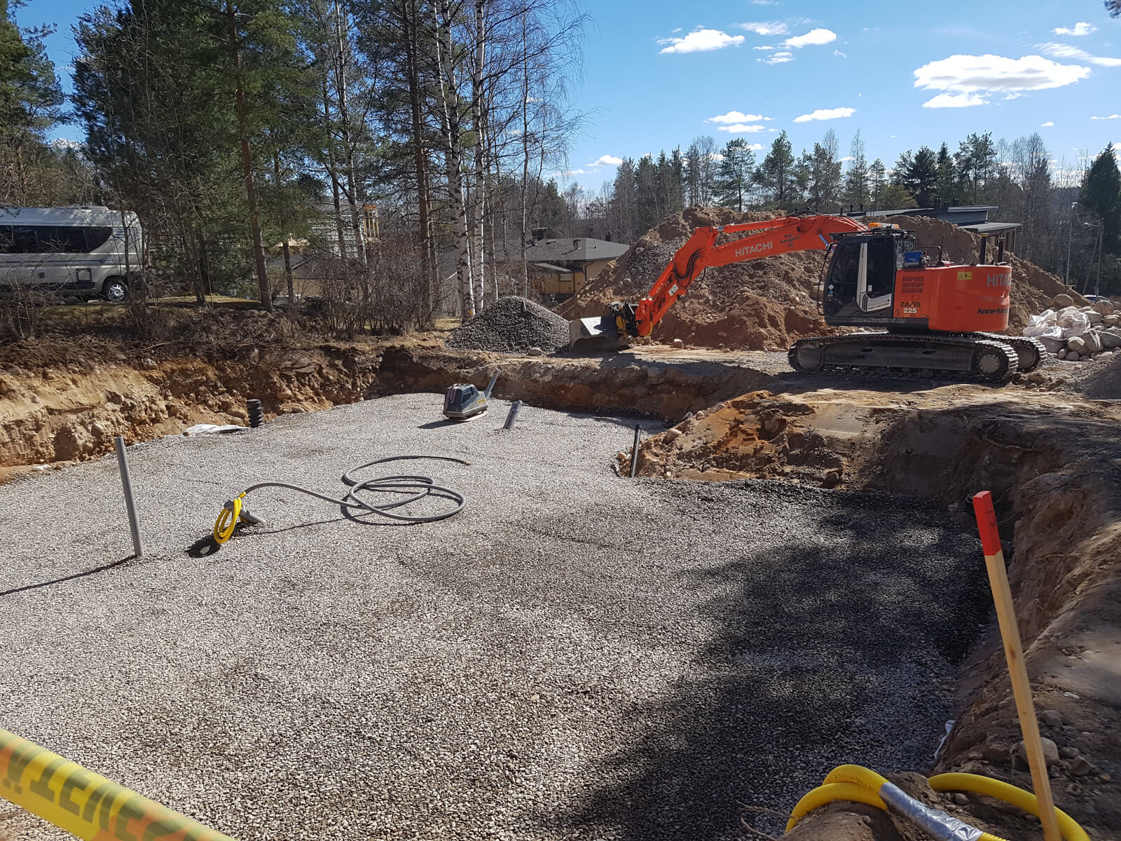 Rakennustyömaa, jossa on oranssi kaivinkone, harmaa asuntovaunu ja kivimurskeella täytetty kuoppa aurinkoisena päivänä.