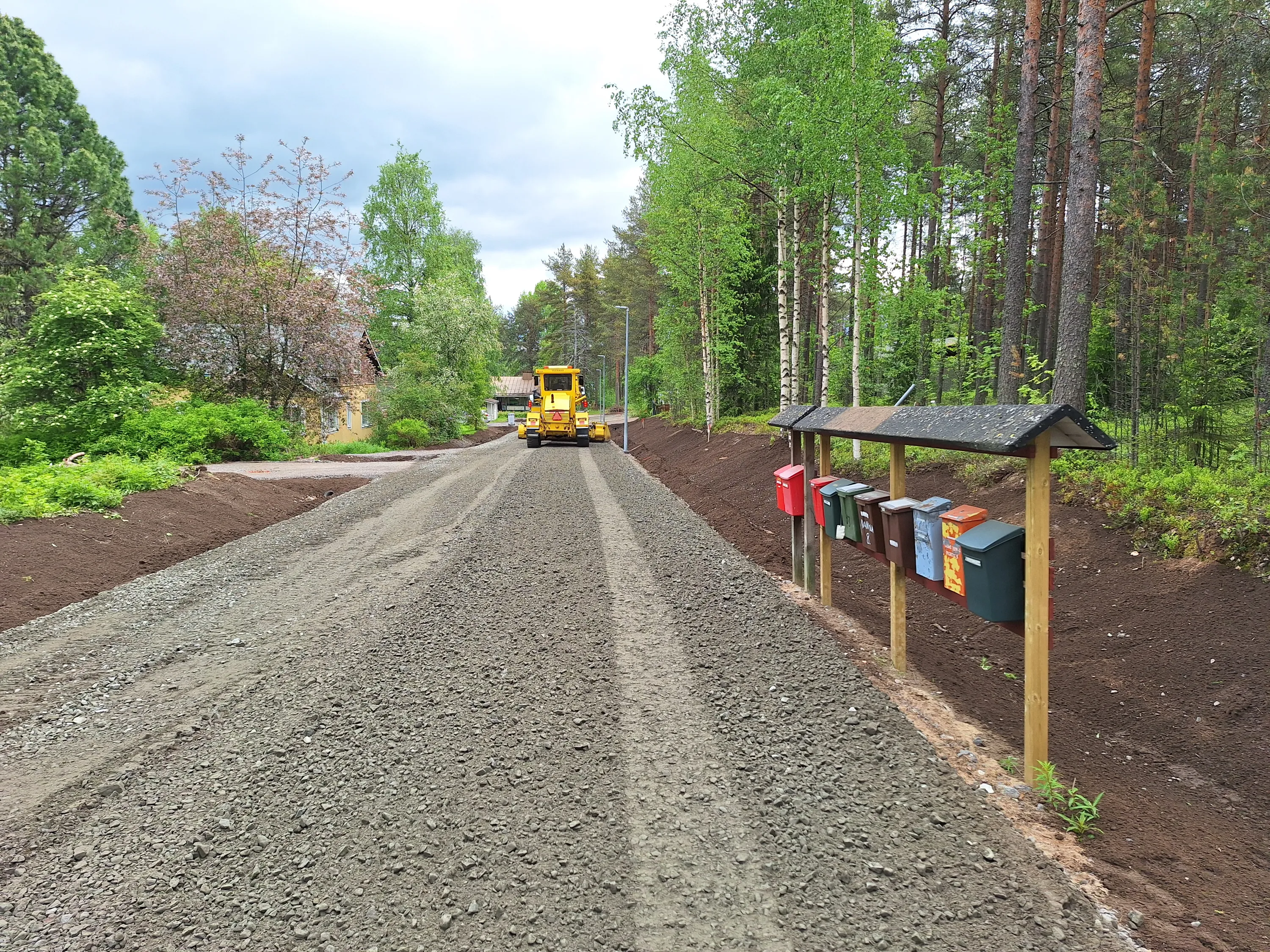 Kuvassa on soratie, jonka varrella on rivi postilaatikoita ja kaukana tiellä näkyy keltainen tiehöylä. Tien molemmin puolin on multaa ja puita.