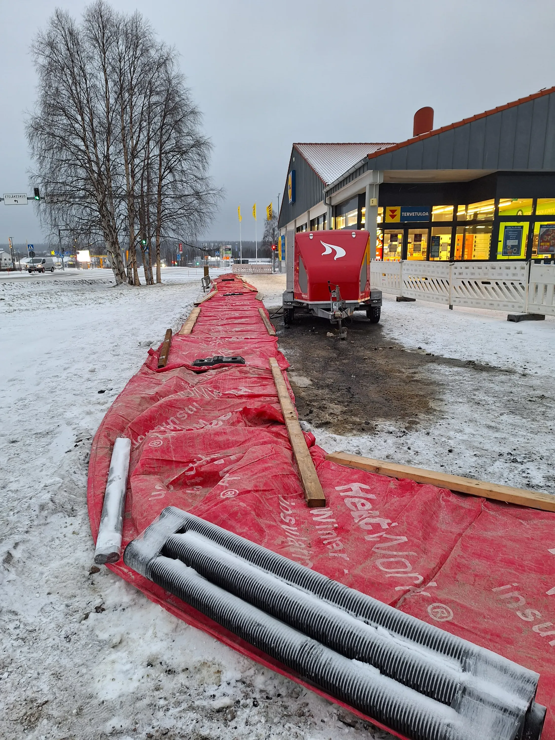 Talvinen maisema, jossa punainen lämmitysmatto, mustat putket, puinen lankku, punainen peräkärry ja Lidl-myymälä taustalla, lumisessa säässä.
