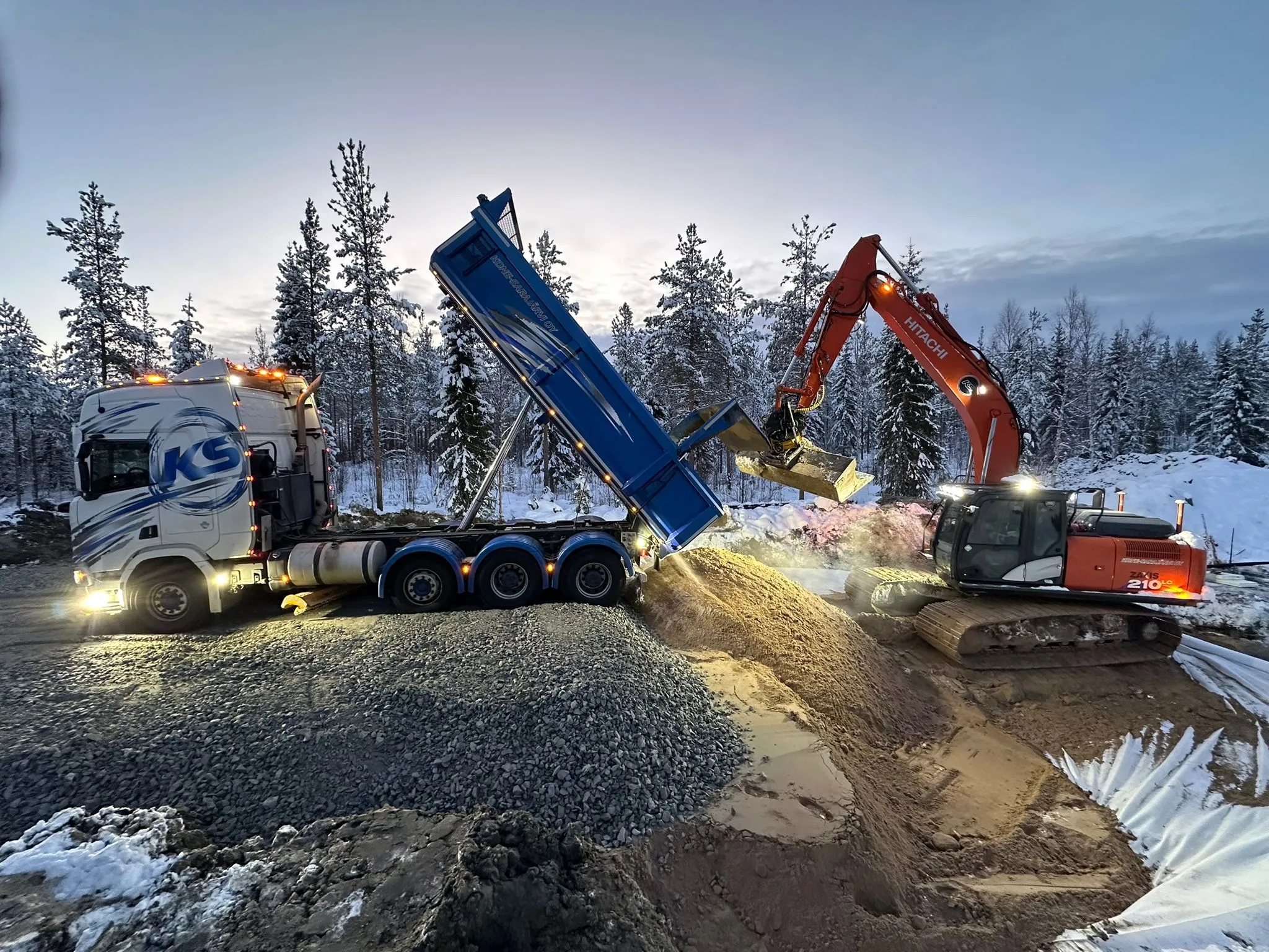 Kuvassa näkyy kuorma-auto, jossa on sininen kippilava, ja oranssi Hitachi-kaivinkone talvisessa maisemassa. Kuorma-auto on valkoinen ja siinä on KS-logo. Maassa on lunta ja soraa.