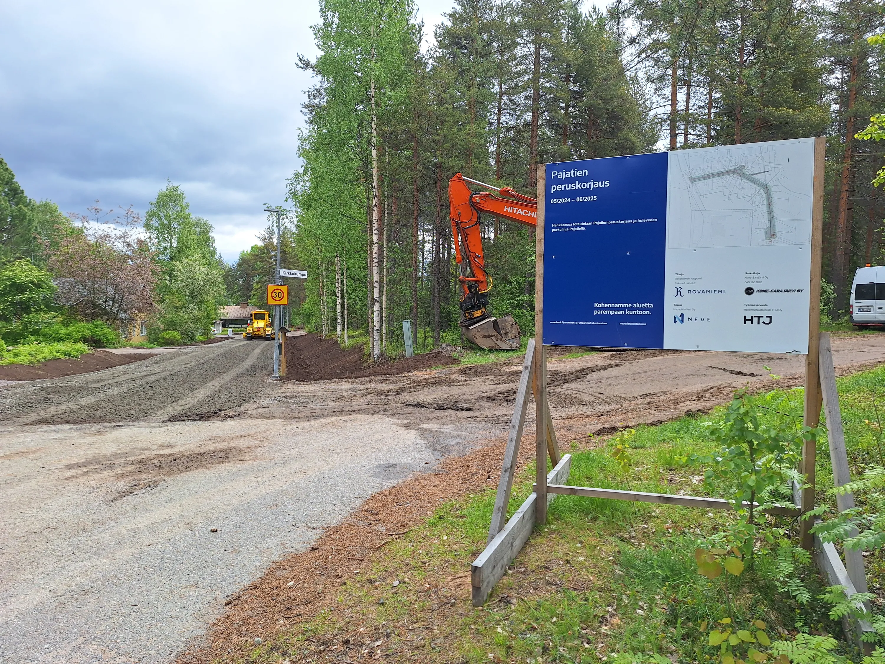 Rakennustyömaa, jossa on tiekunnostustyö, oranssi kaivinkone, keltainen tiehöylä, rakennustyömaan kyltti ja puita.