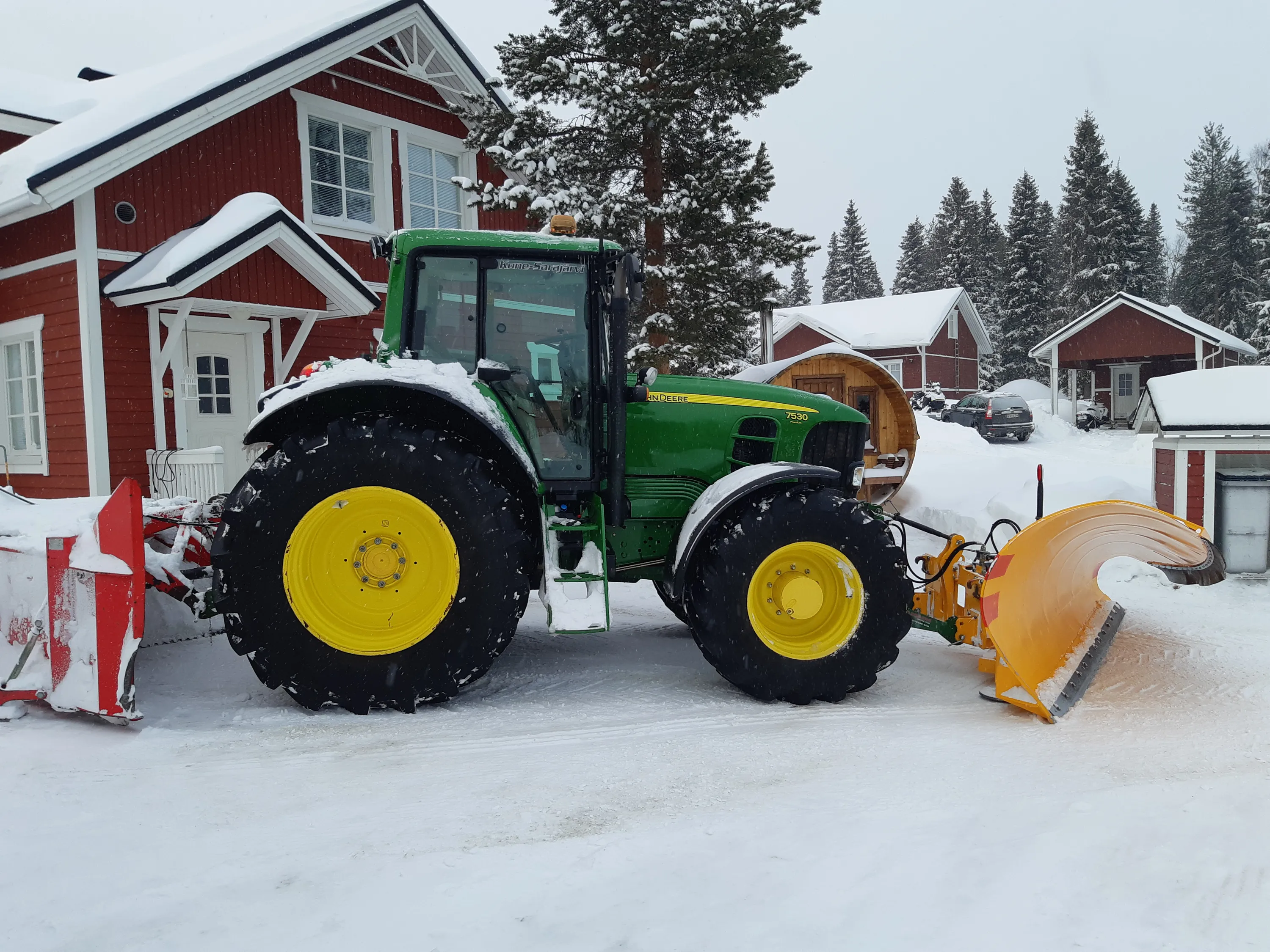 Lumessa seisoo vihreä John Deere 7530 traktori, jossa on keltainen vanteet ja lumiaura edessä ja lumilinko takana. Taustalla on punaisia rakennuksia ja lumisia puita. Valo on himmeä ja sää on luminen.