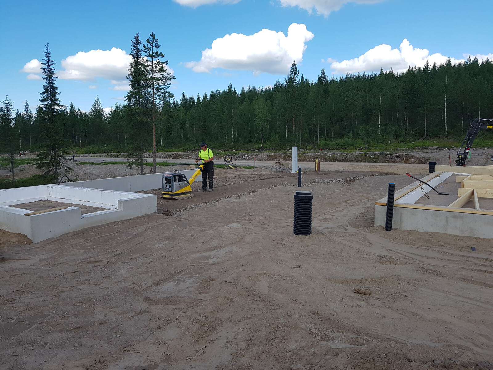 Rakennustyömaalla mies tiivistää maata tärylevyllä, taustalla betonivalu ja metsää, aurinkoinen sää.